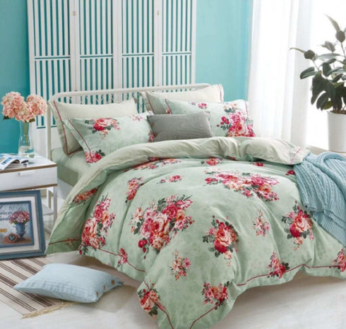 A floral printed top sheet on a bed in a bedroom setting, with various pillows, a plant, and a framed picture beside the bed.