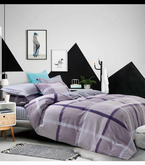 A bed with a purple, gray, and white checkered top sheet, with a single pillow, placed in a room with geometric wall art, a nightstand, and a lamp.