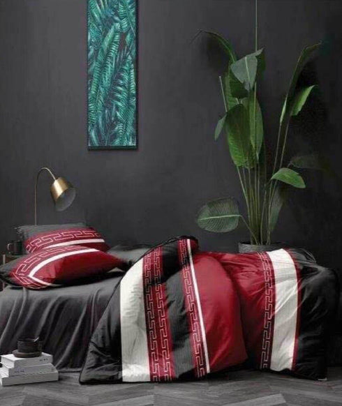 A bed with a multicolored top sheet featuring geometric patterns, with a pillow matching the sheet. The bed is placed against a dark wall with a large painting above it, and there is a lamp and a plant in the room.