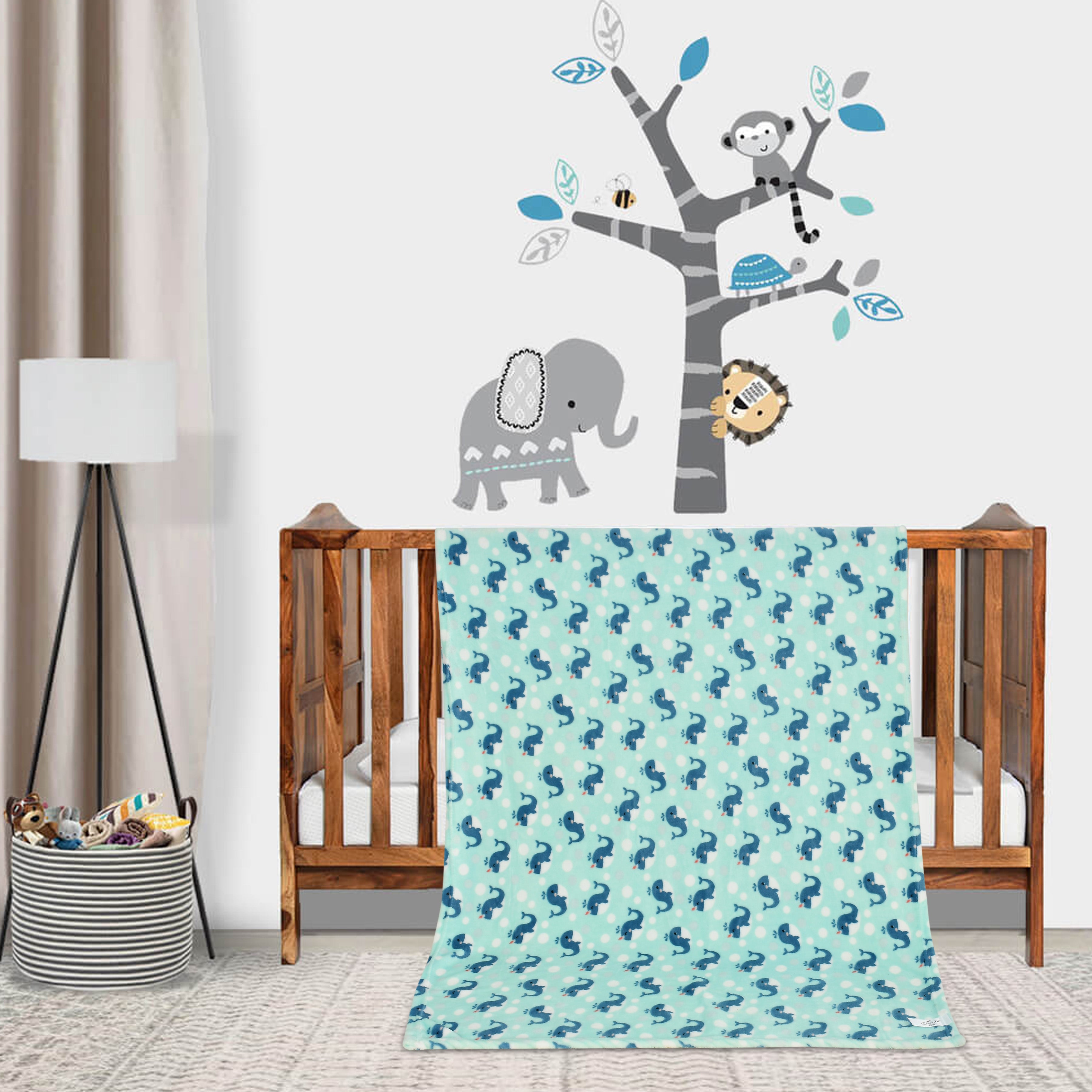 A blue flannel baby blanket with a dolphin print is draped over a crib, with a wall decal featuring a tree with animals and a lamp in the background.