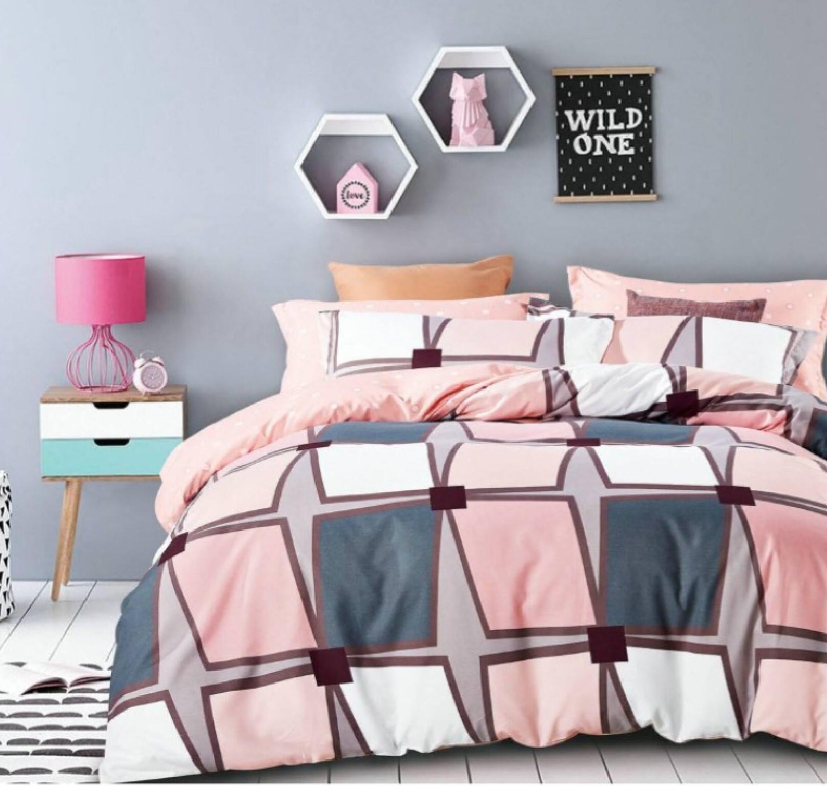 A bedroom scene with a bed having a multicolored geometric patterned top sheet. The room is decorated with pink accents, a nightstand, a lamp, and framed pictures on the wall.